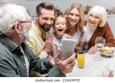 Senior Man Taking Selfie On Mobile Phone With Blurred Family During Easter Dinner