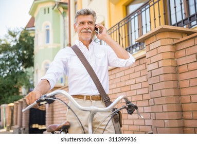 Senior Man Is Talking By Phone Standing With Bike In The Street. Concept Of Active Life Elderly People.