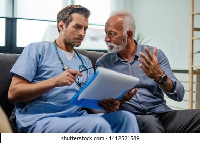 Senior Man Talking To A Doctor Who Is Reading Medical Test Results While Being In A Home Visit. 