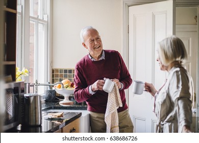 Senior Man Is Talking To His Wife In The Kitchen. He Is Drying Dishes And The Woman Is Dirnking Tea.