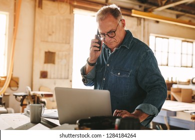 Senior man talking on cell phone and using laptop on work table. Mature carpenter working on laptop and answering phone call in his carpentry workshop. - Powered by Shutterstock