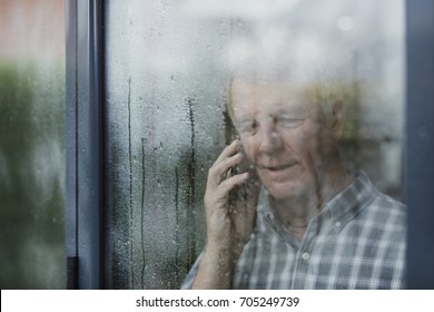 Senior Man Is Talking To Someone On The Phone In His Home. He Is Standing At The Window And It Is Raining Outside. 