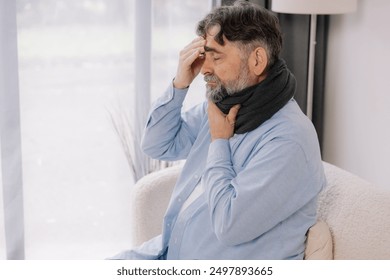 Senior man with throat angina holding his hand to his head sitting on sofa in living room, flu - Powered by Shutterstock