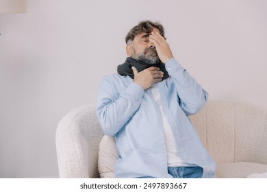 Senior man with throat angina holding his hand to his head sitting on sofa in living room, flu - Powered by Shutterstock