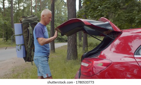 Senior Man Traveler Packing Camping Equipment Into Car Trunk Outdoors. Side View Of Mature Male Hiker Opening Red Car Trunk And Putting Backpack Preparing For Trip