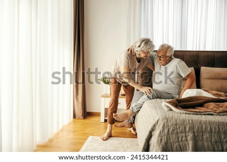 A senior man is trying to get up from a bed in the morning with a help of his wife.