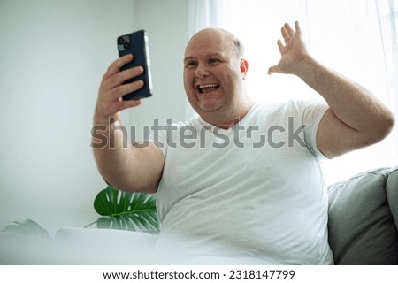 Senior man in t-shirt sweatpants sitdown on grey sofa waving hand talk by video call indoors partment do selfie shot on mobile phone.
