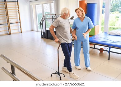 A senior man uses a cane while walking with a physiotherapist in a rehab center for rehabilitation and support after an injury. This indoor physical therapy session focuses on recovery and mobility.