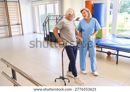 A senior man uses a cane while walking with a physiotherapist in a rehab center for rehabilitation and support after an injury. This indoor physical therapy session focuses on recovery and mobility.