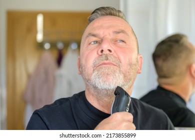 Senior Man Using An Elecrric Trimmer To Trim His Beard In A Bathroom Mirror In A Personal Grooming Concept