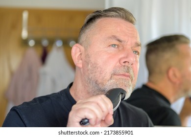 Senior Man Using An Elecrric Trimmer To Trim His Beard In A Bathroom Mirror In A Personal Grooming Concept