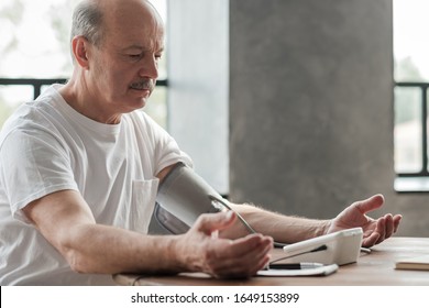 Senior Man Using A Home Blood Pressure Machine To Check His Vital Statistics Sitting At The Living Room