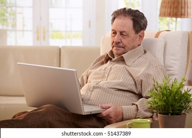 Senior Man Using Laptop Computer At Home, Sitting In Armchair, Looking At Screen.