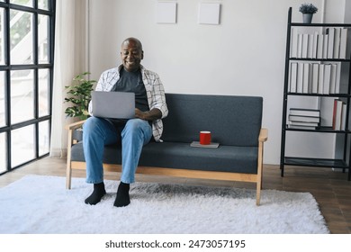 Senior man using laptop on sofa in modern living room with bookshelves and coffee mug, enjoying a relaxed and comfortable environment. - Powered by Shutterstock