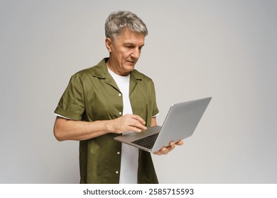 Senior man using laptop while standing against a neutral background with focused expression