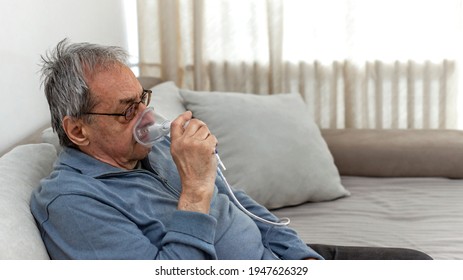 Senior Man Using Medical Equipment For Inhalation With Respiratory Mask. Old Man With Serious Pneumonia Symptoms Caused By Covid-19 Virus Infection Sitting In Home During Lockdown, Wearing Oxygen Mask