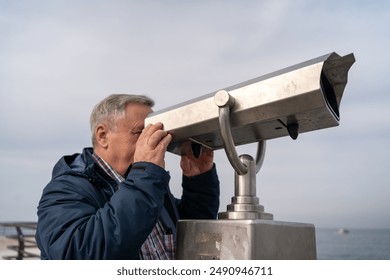 Senior man using public telescope at seaside. Elderly gentleman observing distant view through coin-operated binoculars. - Powered by Shutterstock