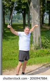 Senior Man Using Rings To Work Out At An Outdoor Sports Facility In A Lush Green Park Standing Looking At The Camera With A Smile In A Health And Fitness Concept