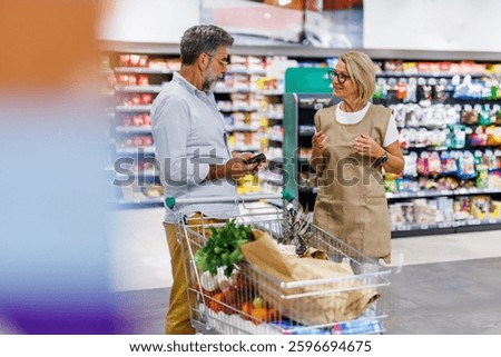 Senior man using a smartphone and talking to a supermarket employee, standing next to a shopping cart full of groceries inside a supermarket