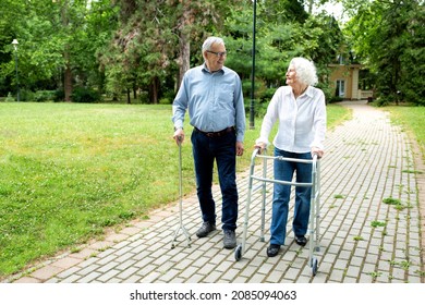 Senior Man Using A Walking Cane Accompanied By A Senior Lady Strolling With Folding Walker In The Park