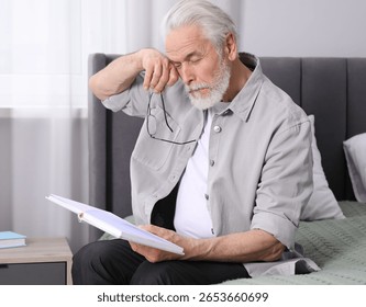 Senior man with vision problem reading book on bed at home