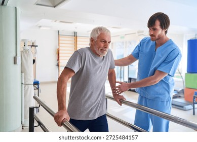 A senior man is walking with parallel bars assisted by a physiotherapist in a rehab center. This image can be used commercially for injury recovery mobility training and healthcare related concepts indoors.