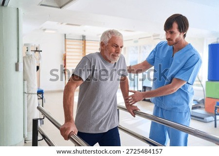 A senior man is walking with parallel bars assisted by a physiotherapist in a rehab center. This image can be used commercially for injury recovery mobility training and healthcare related concepts indoors.