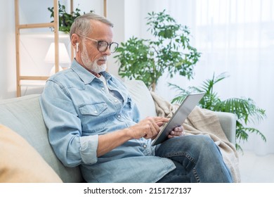 Senior man watches morning breaking news on tablet sitting on sofa against lush pot-plant. Elderly bearded gentleman in glasses uses gadget at home. work from home during lockdown