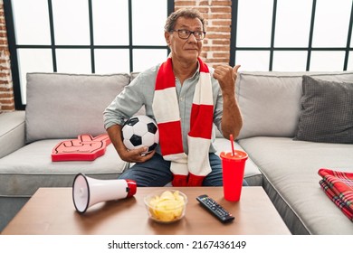 Senior man watching football holding ball supporting team pointing thumb up to the side smiling happy with open mouth 