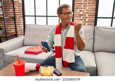 Senior Man Watching Football Looking At Smartphone Pointing Thumb Up To The Side Smiling Happy With Open Mouth 