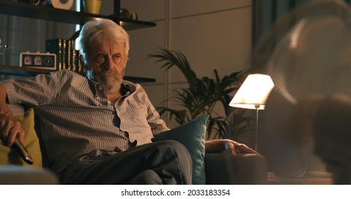 Senior Man Watching TV At Home On A Summer Night, Fan In The Foreground