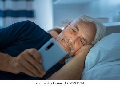 Senior Man Watching Video On Smart Phone While In Bed. Smiling Mature Man Lying On Bed Using Cellphone In The Dark At Home. Addict Man Awake Using Phone For Chatting Late In Night.