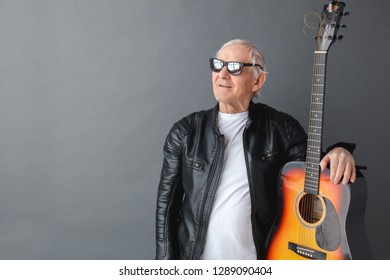 Senior Man Wearing Black Leather Jacket And Sunglasses Standing Isolated On Gray Wall Hugging Guitar Looking Aside Dreamful