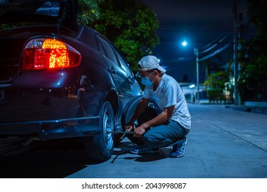 Senior Man Wearing A Mask Change Tire Roadside At Night.Accident, Broken Car, Broken Tire, Waiting For Help.