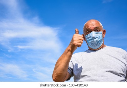 Senior Man Wearing Medical Mask And Goggles Looking At The Camera And Shows Thumbs Up. Old Man On A Blue Sky With Clouds Background