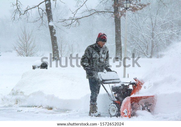 Senior man well dressed for a\
Chicago blizzard in wool stocking cap, canvas jacket and ear\
protection snow blowing his Driveway during a Chicago winter\
storm.