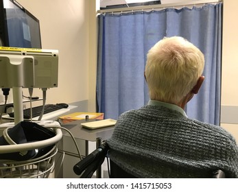 Senior Man In A Wheelchair, Waiting To Be Assessed On The Accident And Emergency Ward In A Hospital.  Man Viewed From Behind, In The Cubicle With Curtain Drawn.