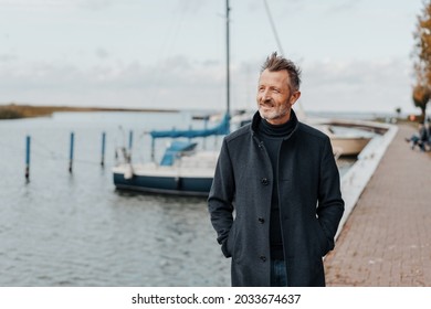 Senior Mann in einem WinterüberMantel, der an einer Promenade entlang einer Marina mit Yachten an einem kalten Tag in einem gesunden, aktiven Outdoor-Lifestyle-Konzept spaziert