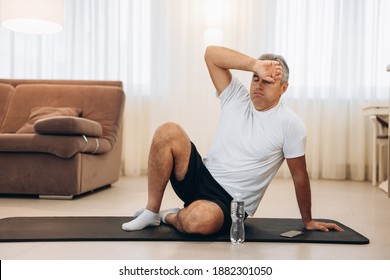 Senior Man Wipes Sweat Of His Forehead After Hard Home Workout. Healthy Lifestyle Concept. Living Room On Background. Man Tired After Intensive Training. Plastic Water Bottle Near Black Yoga Mat.