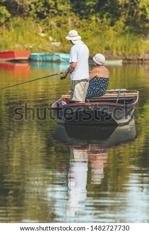 Senior man and woman are fishing from a wooden boat in the center of a freshwater a lake fringed with lush scenery.