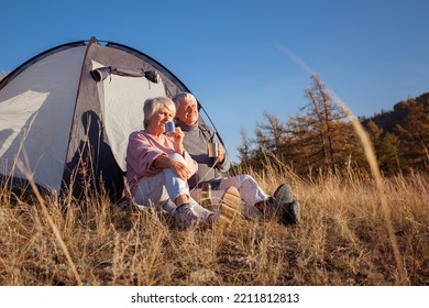 Senior Man And Woman Looks Into The Distance Near The Tent At The Mountain, Adult Couple Sitting At Camping Site At Autumn Fall Season