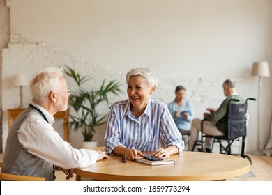 Senior man and woman sitting at table and enjoying talk, another aged couple interacting in background sitting on sofa in common room of nursing home - Powered by Shutterstock