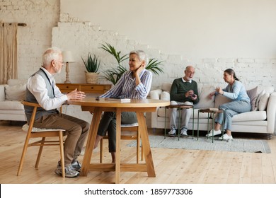 Senior man and woman sitting at table and enjoying talk, another aged couple interacting in background sitting on sofa in common room of nursing home - Powered by Shutterstock