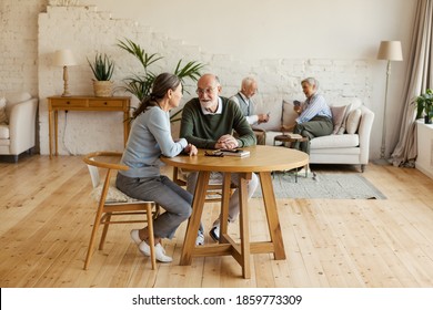 Senior Man And Woman Sitting At Table And Talking, Another Aged Couple Playing Cards In Background Sitting On Sofa In Common Room Of Assisted Living Home