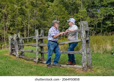 Senior Man and Woman talking with each other, leaning over an old country rail fence along a field of grass and trees.  Start of autumn green turning to gold and brown. Blue jeans and ballcaps. 