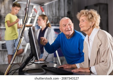 Senior Man And Woman Using Computer To Solve Conundrum In Escape Room. Their Grandson Standing On Stepladder Behind Them And Giving Metal Can To Teenager Girl.