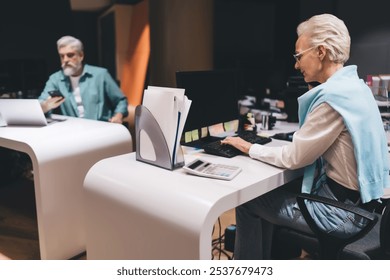 Senior man and woman working at separate desks in a modern office. Woman on phone using computer and calculator, while man checks his smartphone beside a laptop, symbolizing multitasking and focus - Powered by Shutterstock