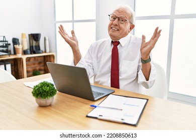 Senior Man Working At The Office Using Computer Laptop Clueless And Confused Expression With Arms And Hands Raised. Doubt Concept. 