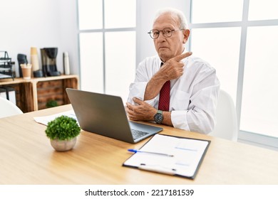 Senior Man Working At The Office Using Computer Laptop Pointing With Hand Finger To The Side Showing Advertisement, Serious And Calm Face 