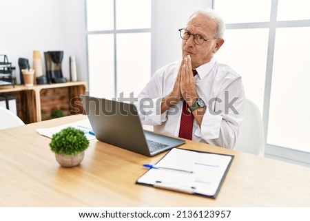 Senior man working at the office using computer laptop praying with hands together asking for forgiveness smiling confident. 